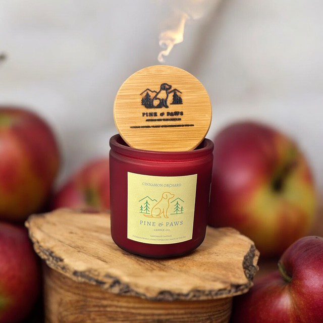 A red apple-scented candle with wooden lid on a wooden slice, surrounded by apples.