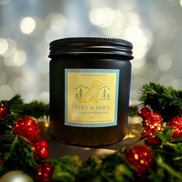 A lit scented candle in a dark jar surrounded by festive holiday decorations.