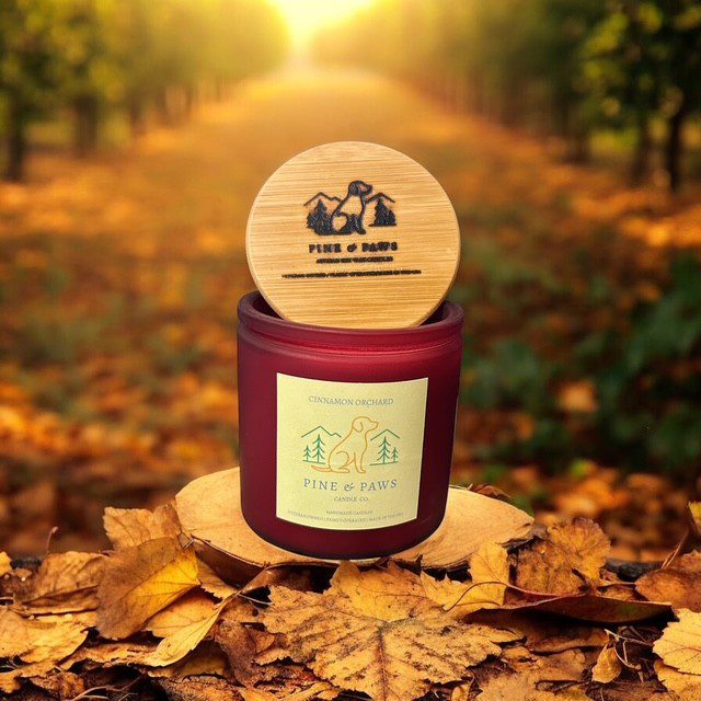 A candle jar with a wooden lid sits on autumn leaves in a sunlit orchard.