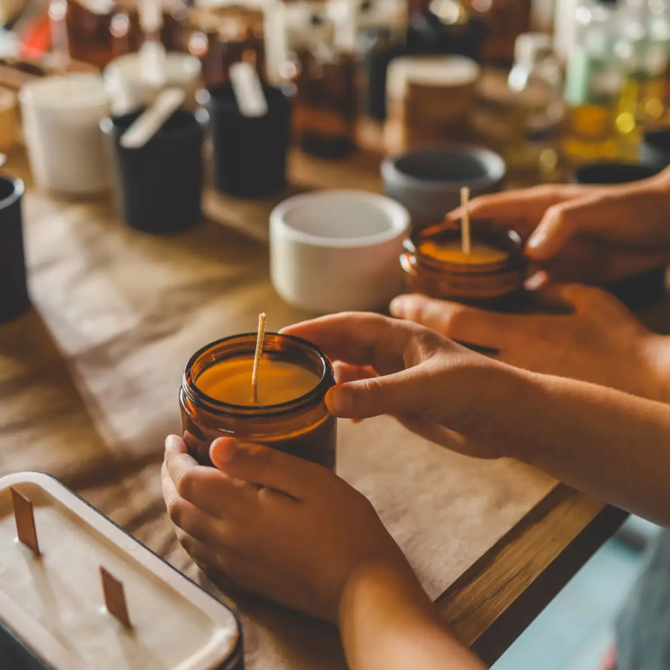 Hands stirring a candle jar on a wooden table with other candle supplies.